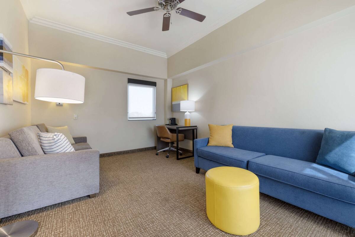 A modern living room with a grey sofa, a blue sofa, a yellow ottoman, a desk with a chair, and a ceiling fan.