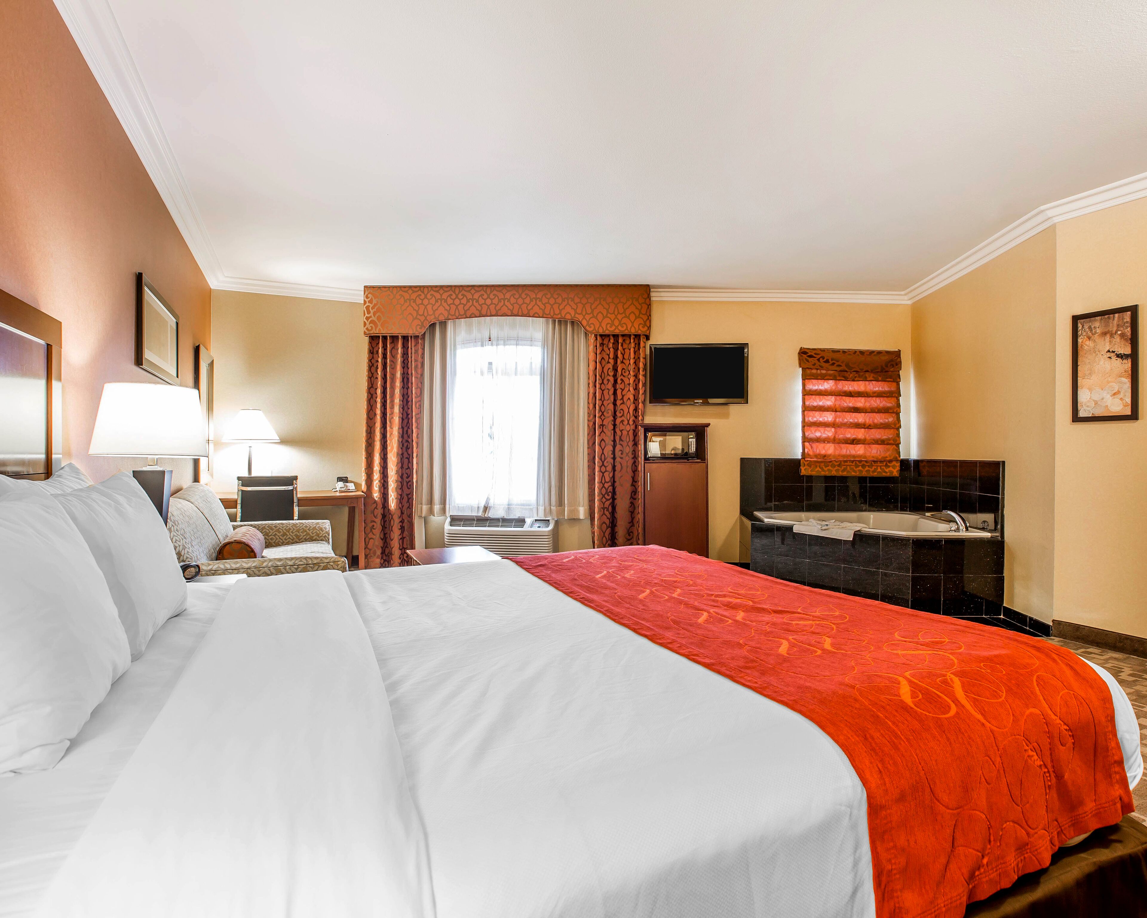 A hotel room with a large bed, a desk with a lamp, a television, a bathroom with a bathtub, and a window with curtains.