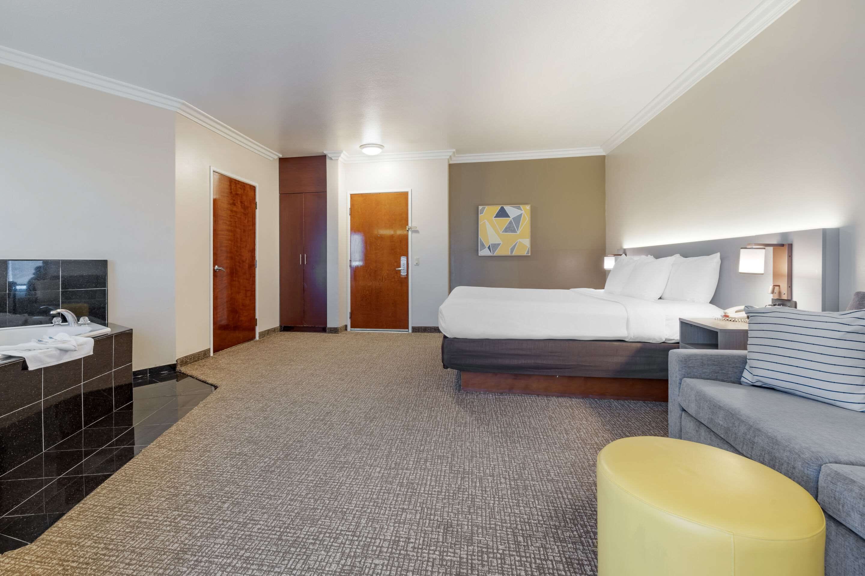 A hotel room with a bed, a sofa, a small table with a lamp, a bathroom with a sink and mirror, and a door leading to another room.