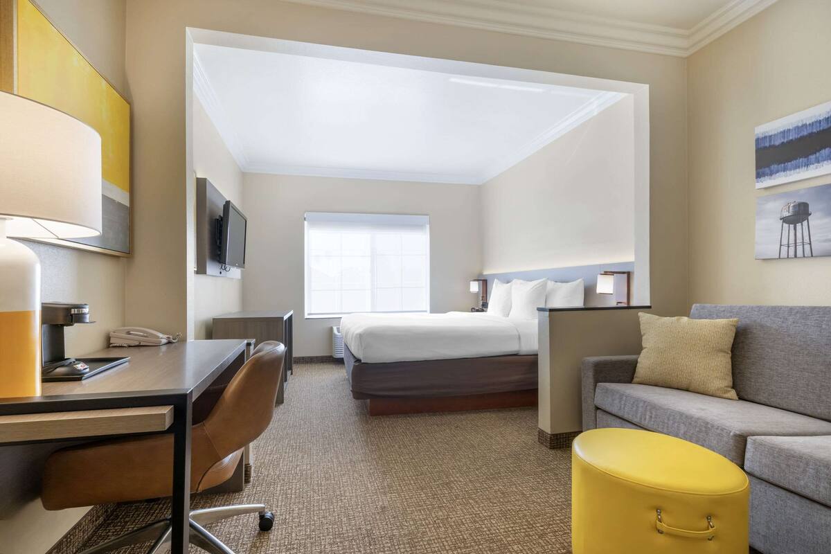 A hotel room with a bed, a desk, a chair, a sofa, a small yellow ottoman, a TV, and a window.