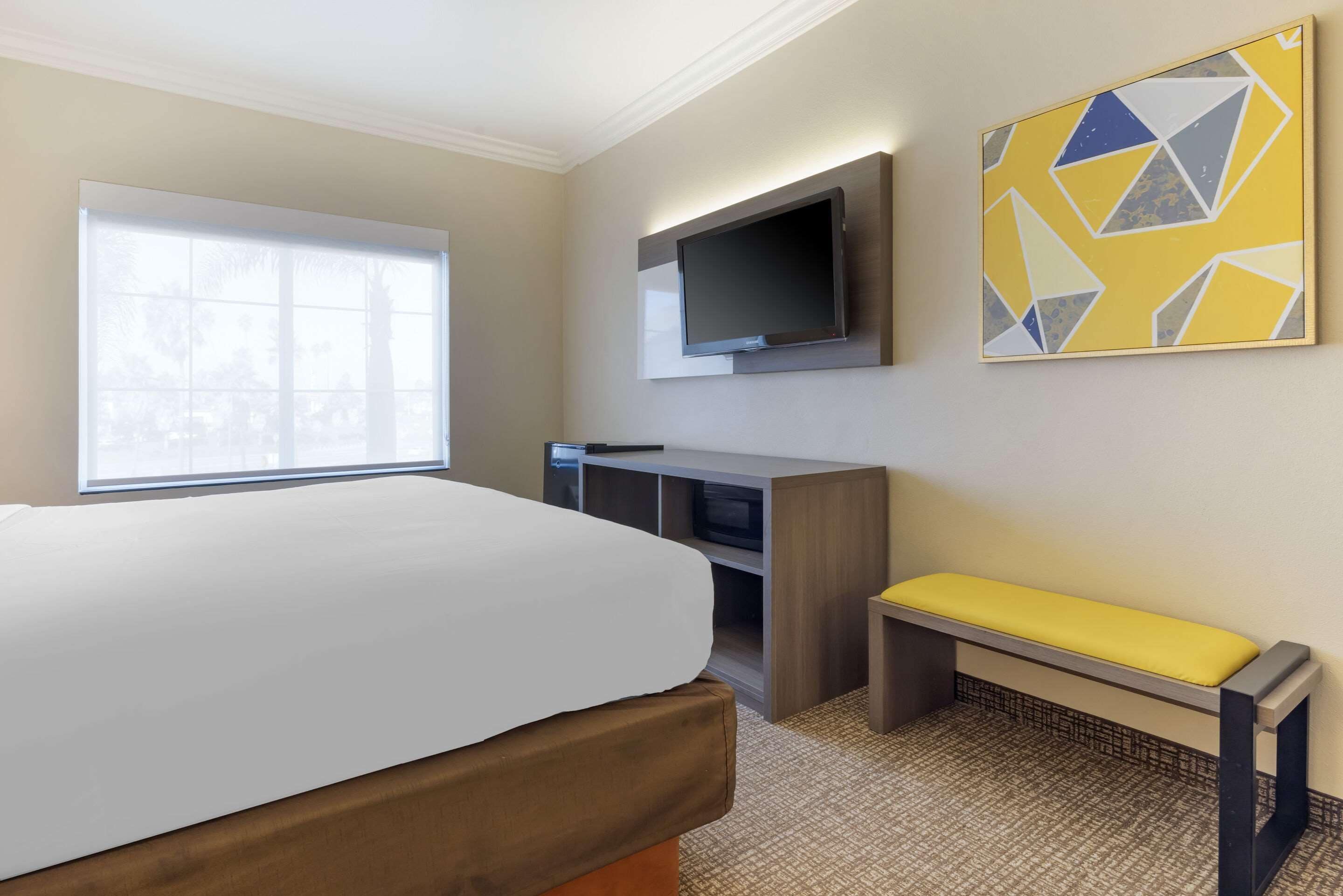 Una habitación de hotel con una cama grande, un escritorio con televisor de pantalla plana, un banco y una ventana con persianas.