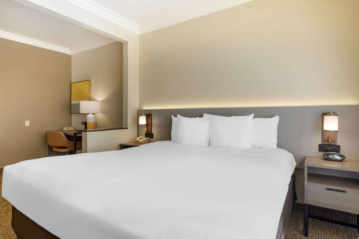 A hotel room with a large bed, bedside tables, a desk with a chair, and a lamp.