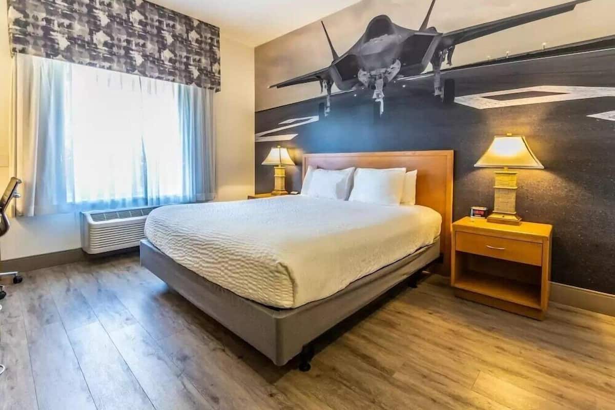 Une chambre d’hôtel comprenant un lit, une table de chevet, une lampe, une œuvre d’art accrochée au mur et une fenêtre avec des rideaux.