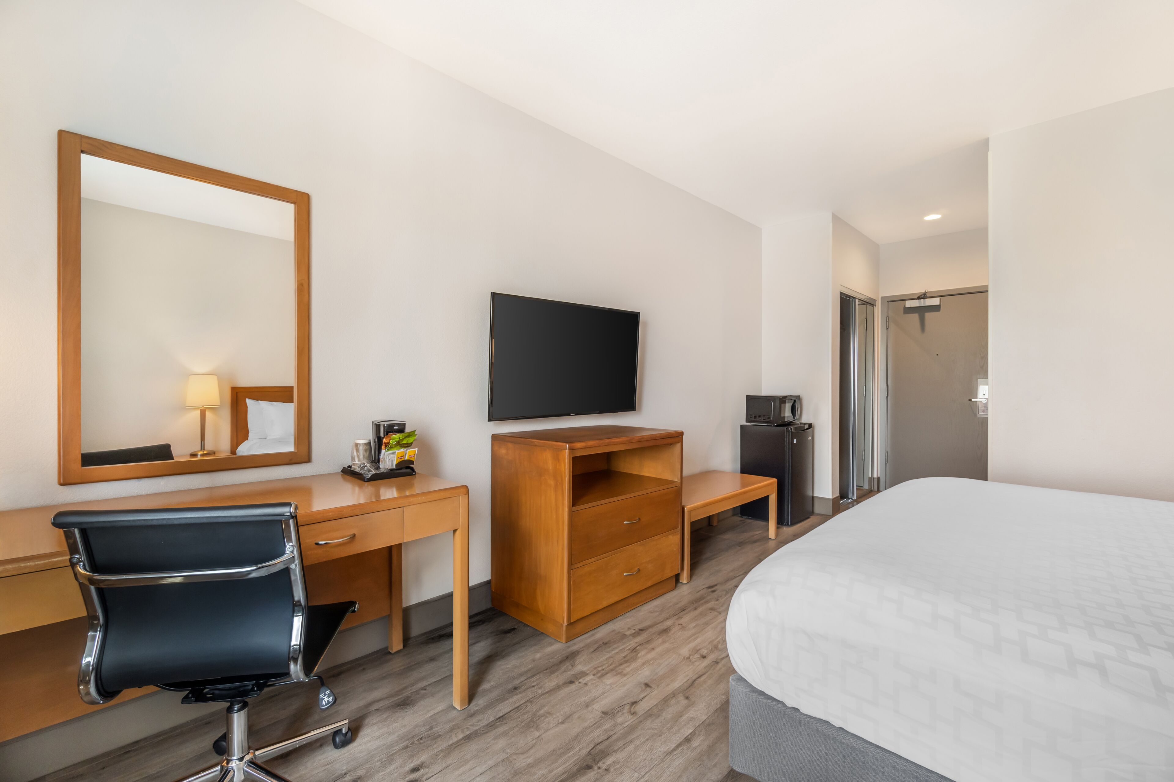 Ett hotellrum med en säng, ett skrivbord, en stol, en tv, en spegel och en dörr.