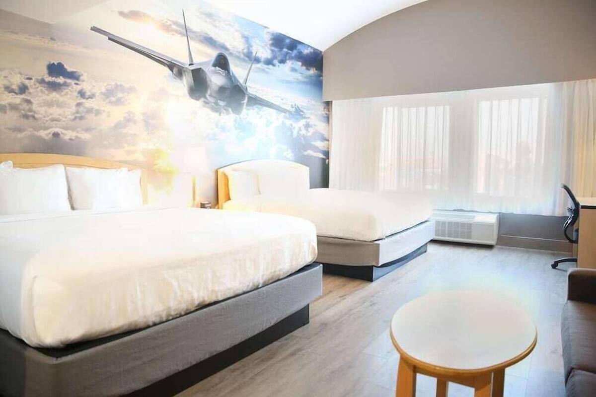 Une chambre d’hôtel avec un grand lit, un lit plus petit, une table ronde et une fresque représentant un avion.