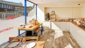 Un área de desayuno con un sistema tipo bufé, que incluye una máquina de café, varios recipientes con alimentos y un espacio de estar con mesas y sillas.