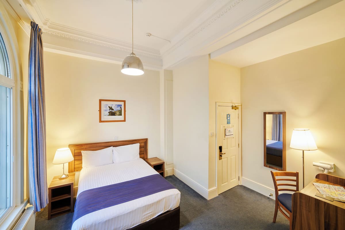 A hotel room with a bed, bedside tables, a desk, a chair, a desk lamp, a window with curtains, and a door.