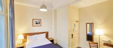 A hotel room with a bed, bedside tables, a desk, a chair, a desk lamp, a window with curtains, and a door.