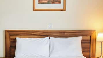 A neatly made bed with white linens and a blue accent, a framed picture of a building on the wall, and a bedside lamp.