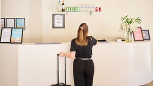 Un mostrador de recepción con una mujer detrás, sosteniendo una maleta. Hay diplomas enmarcados en la pared, un estante con bebidas y un jarrón con flores sobre el mostrador.