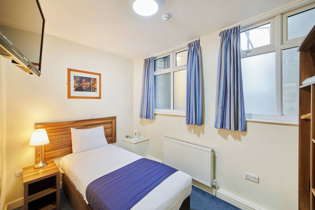 A hotel room with a bed, bedside table, lamp, window with curtains, and a picture on the wall.