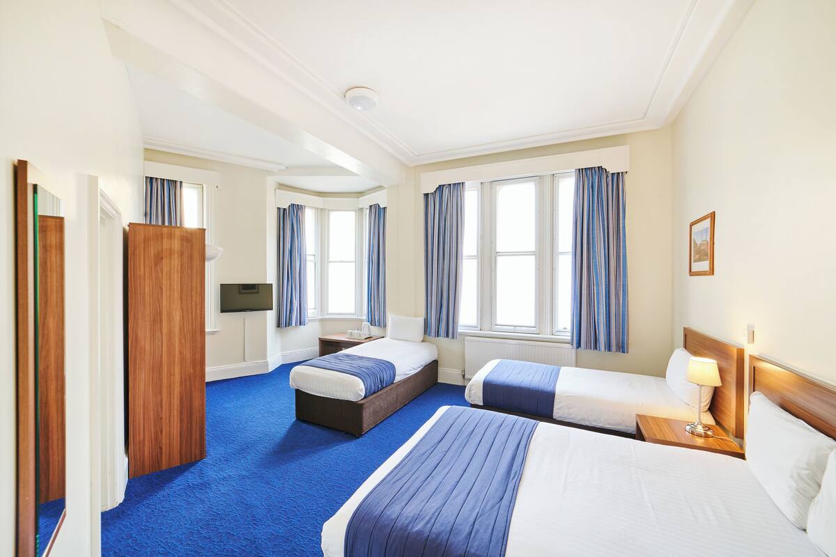 A hotel room with two beds, a wooden wardrobe, a television, and large windows with curtains.