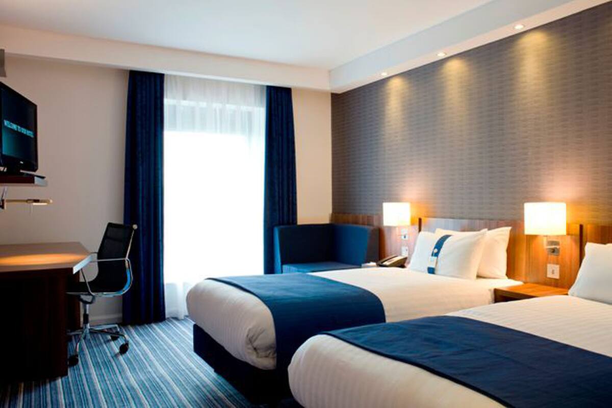 A hotel room with two beds, a desk, a chair, a television, and a large window with curtains.