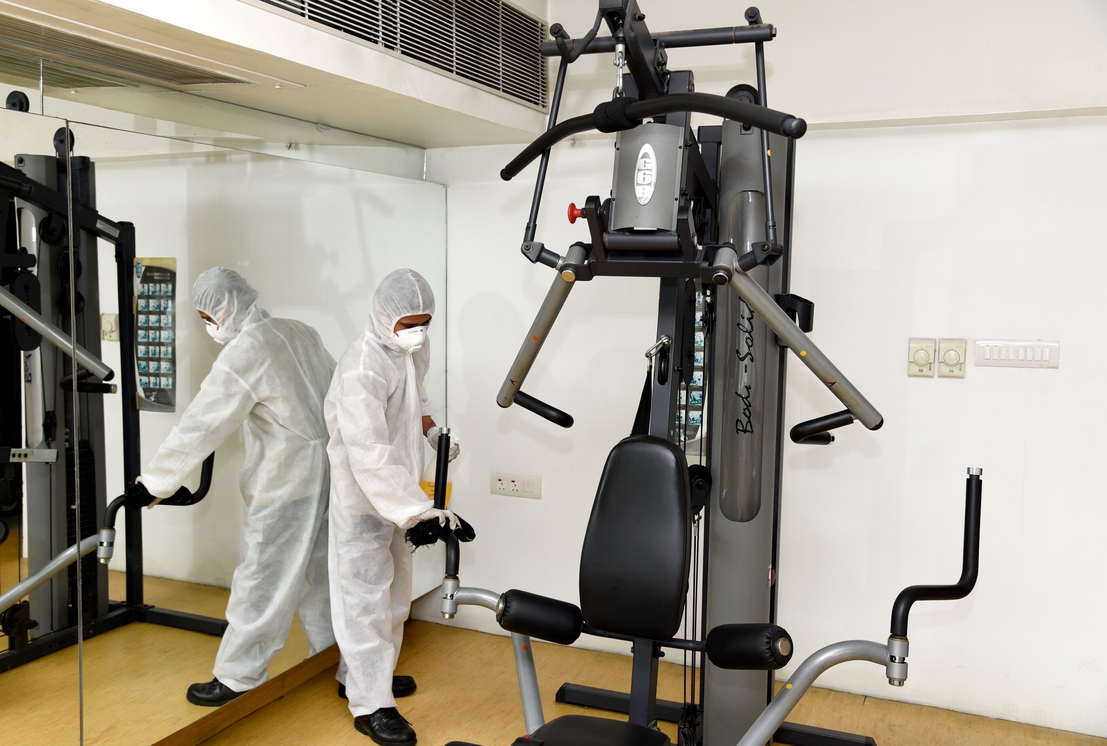 Two individuals in protective suits using exercise equipment in a room with a white wall and a vent.