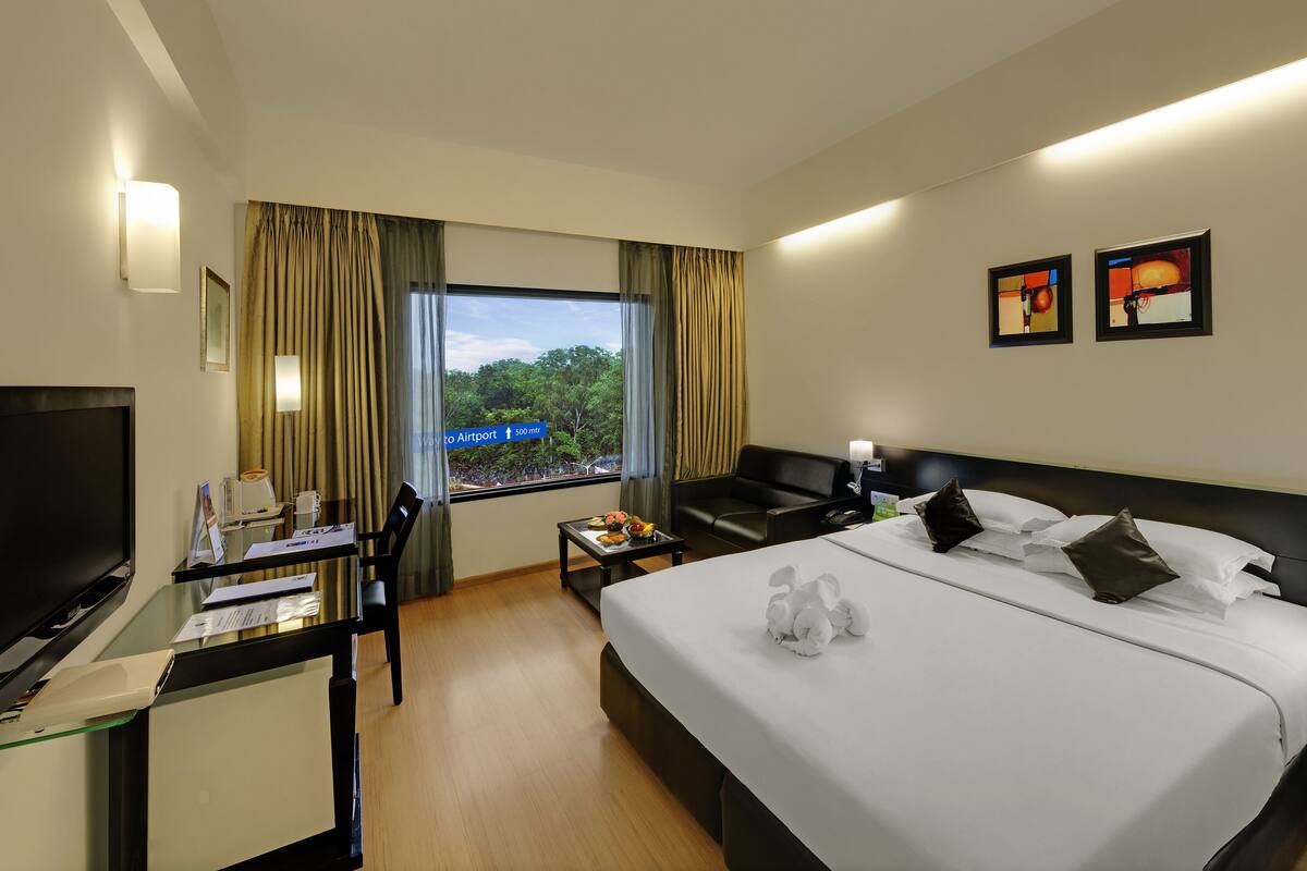 A hotel room with a bed, bedside table, TV, desk with computer, and a window with curtains.