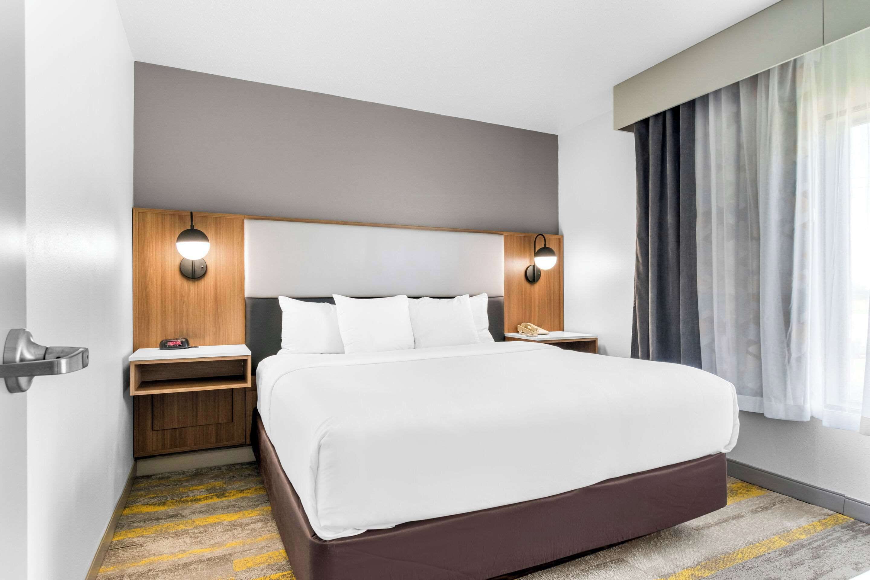 A modern hotel room with a large bed, wooden headboard, two bedside tables with lamps, a telephone, and a window with curtains.