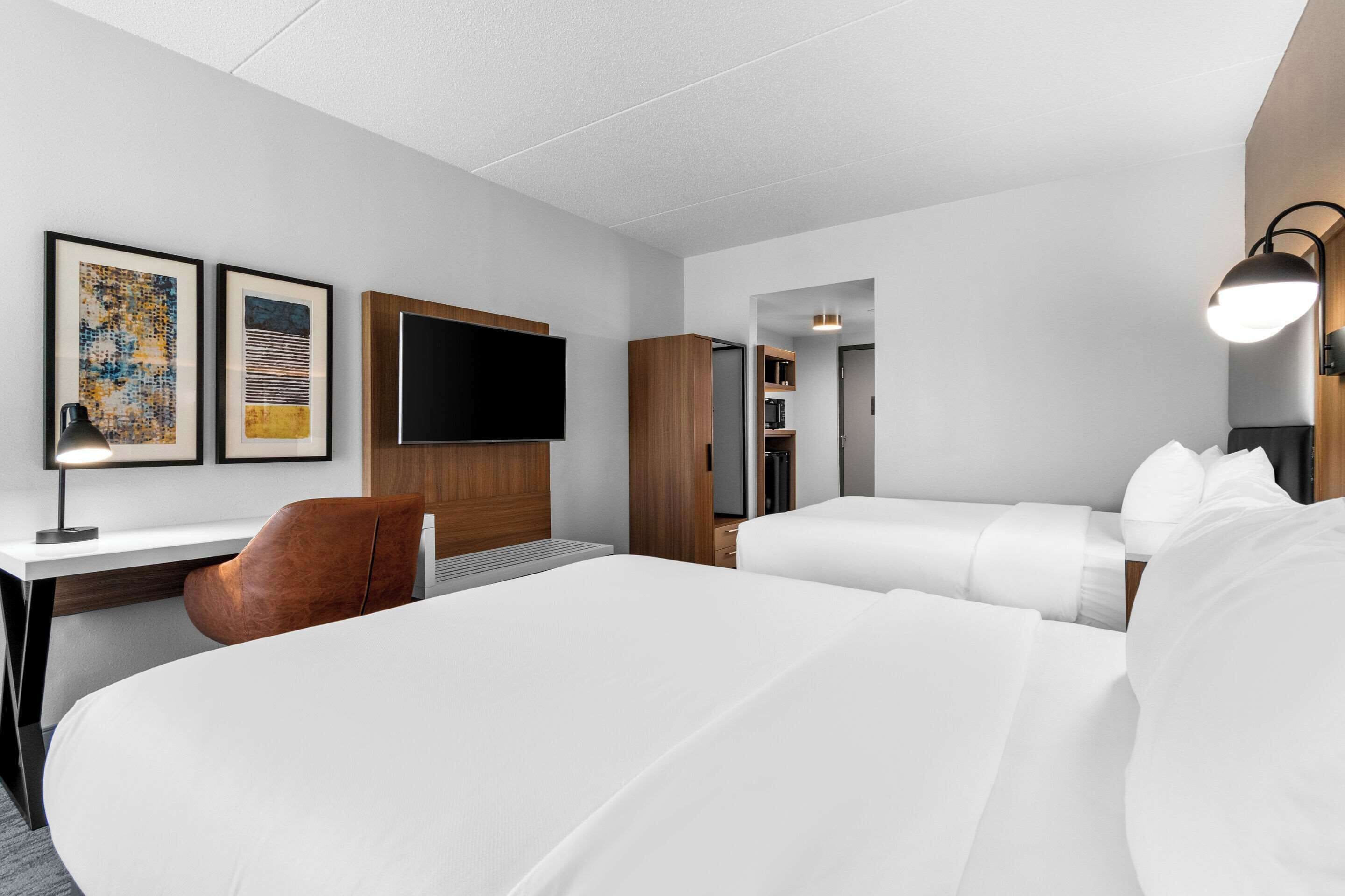A hotel room with two beds, a desk, a chair, a flat-screen TV, and artwork on the wall.