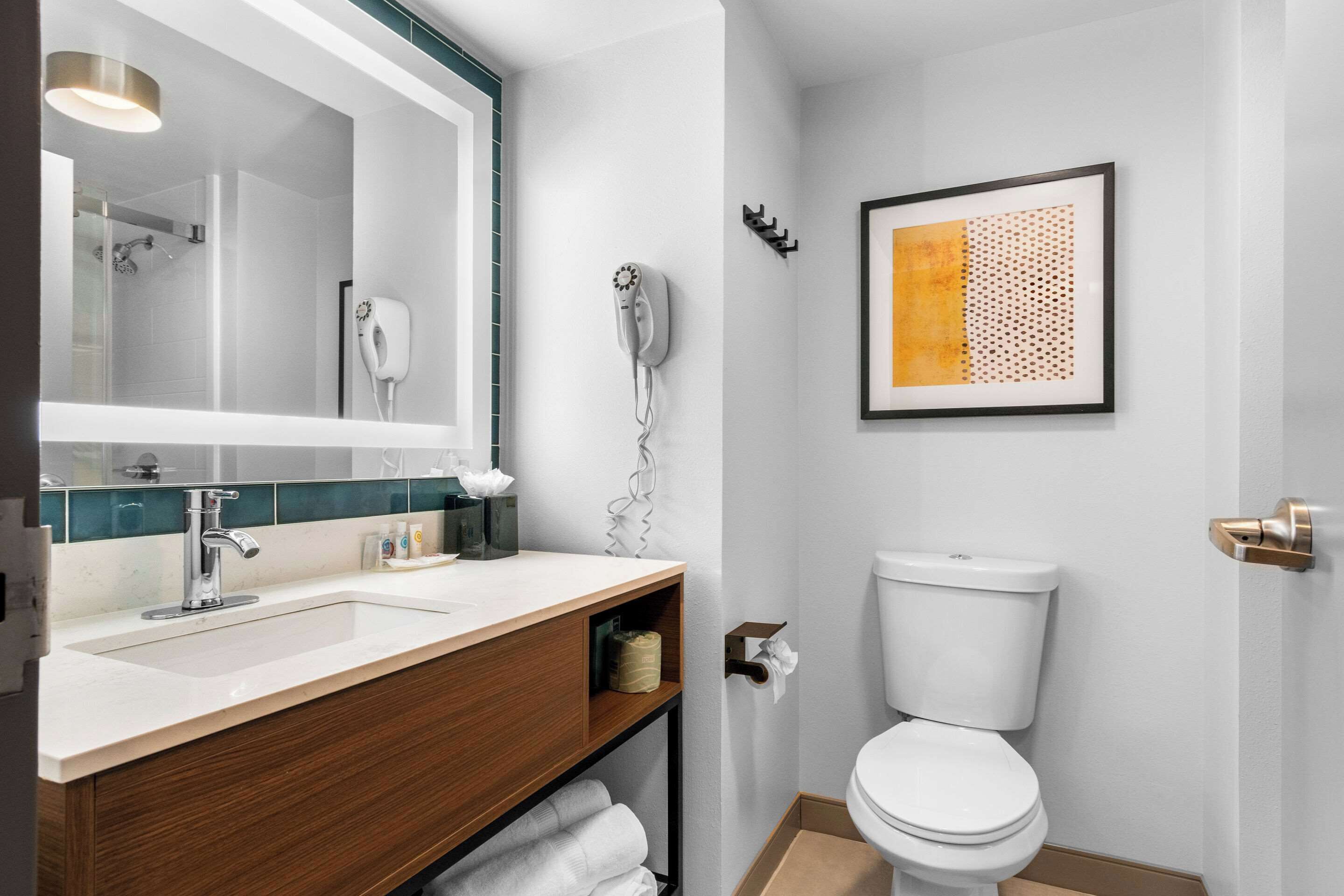 A modern bathroom with a white sink, wooden vanity, a large mirror, a toilet, and a framed artwork on the wall.