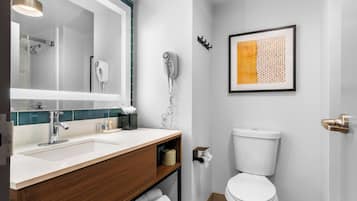 A modern bathroom with a white sink, wooden vanity, a large mirror, a toilet, and a framed artwork on the wall.