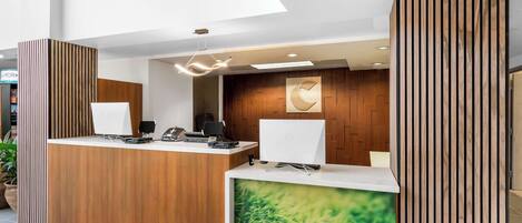 Um balcão de recepção moderno com acabamento em madeira, dois monitores de computador, um telefone e uma tela grande exibindo uma paisagem natural.