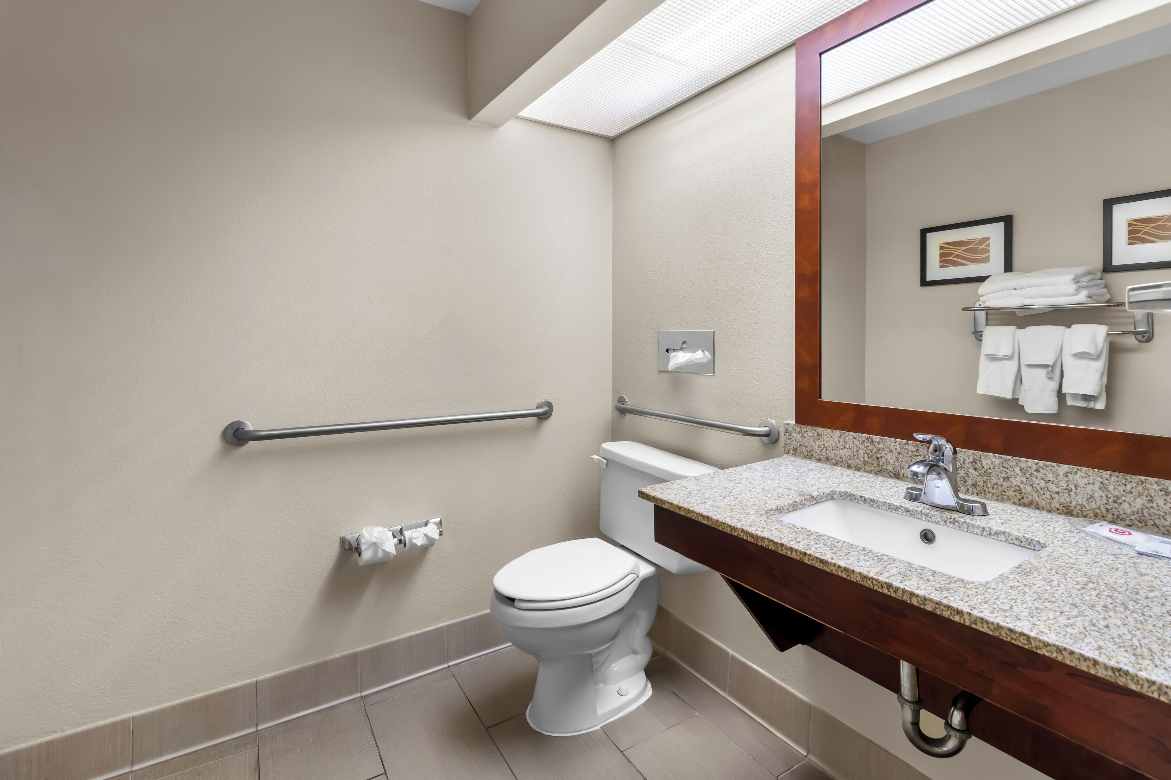 Une salle de bain moderne avec des toilettes blanches, un lavabo doté d’un plan de travail en granit et un miroir au-dessus du lavabo. Des barres d’appui sont fixées aux murs, et un porte-serviettes contient des serviettes pliées.