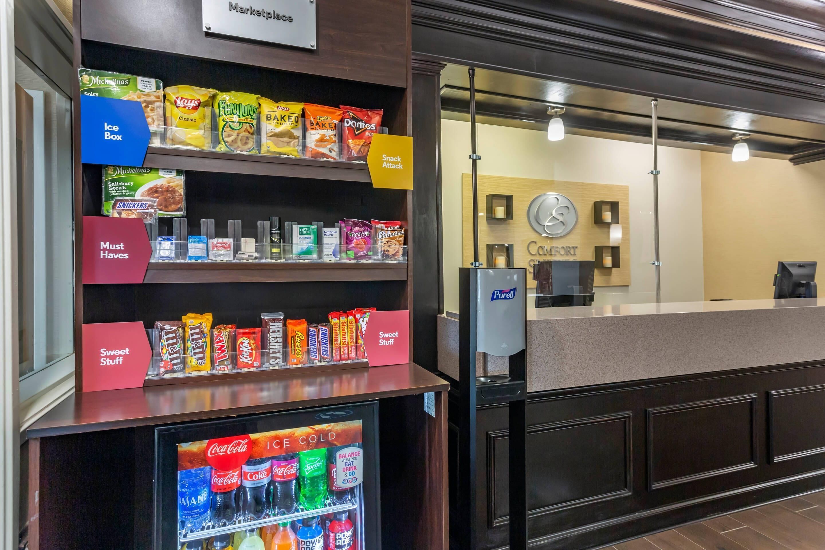 A hotel reception area with snack and ice box shelves, a refrigerator with beverages, and a counter with a clock and a sign reading "Comfort".
