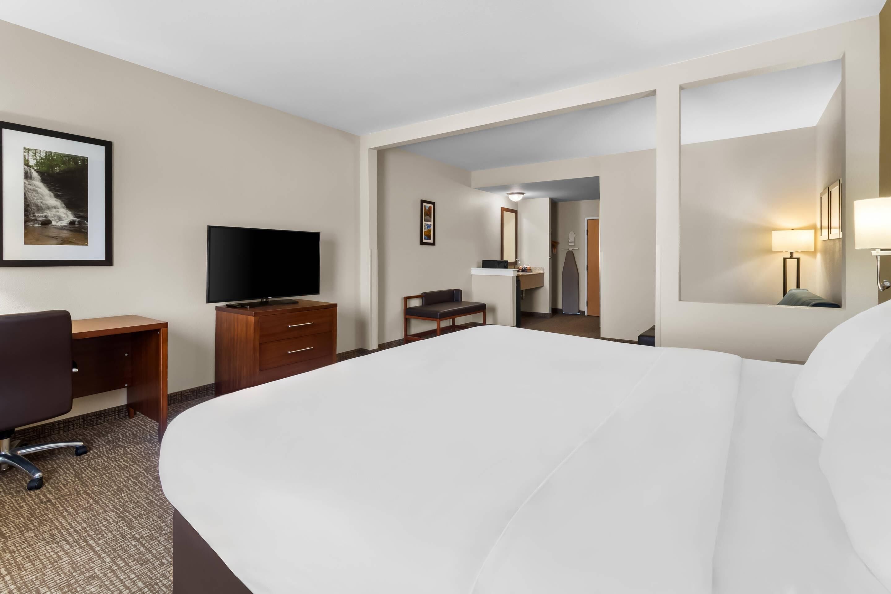A hotel room with a large bed, a desk with a TV, a chair, a wardrobe, and a bathroom visible through an open door.