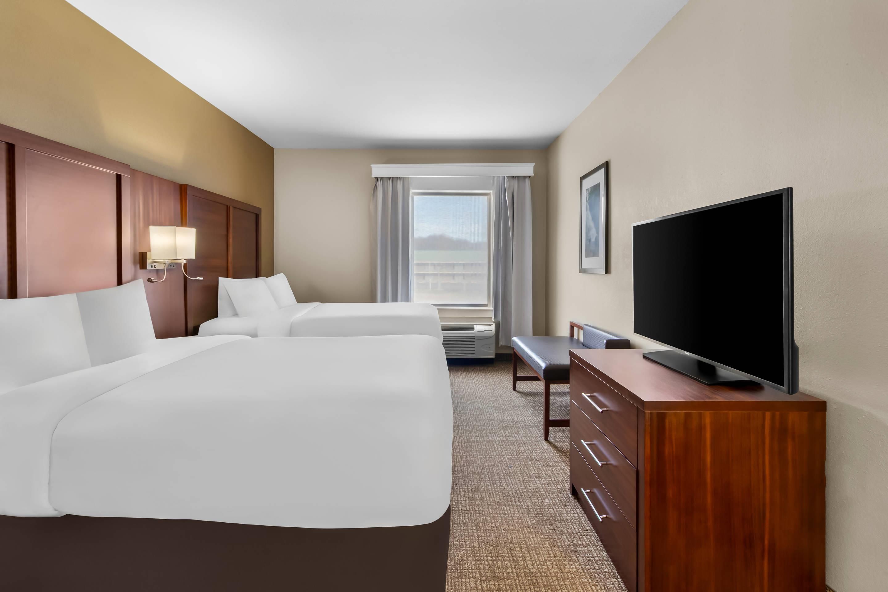 Una habitación de hotel con una cama grande, un televisor sobre un tocador de madera y vistas al exterior a través de la ventana.