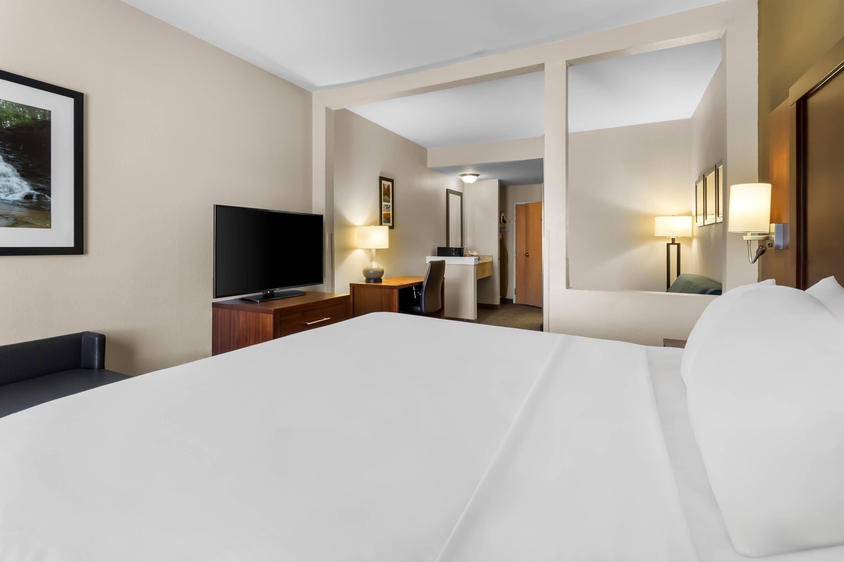 A hotel room with a large bed, a flat-screen TV, a desk with a lamp, and a door leading to another room.
