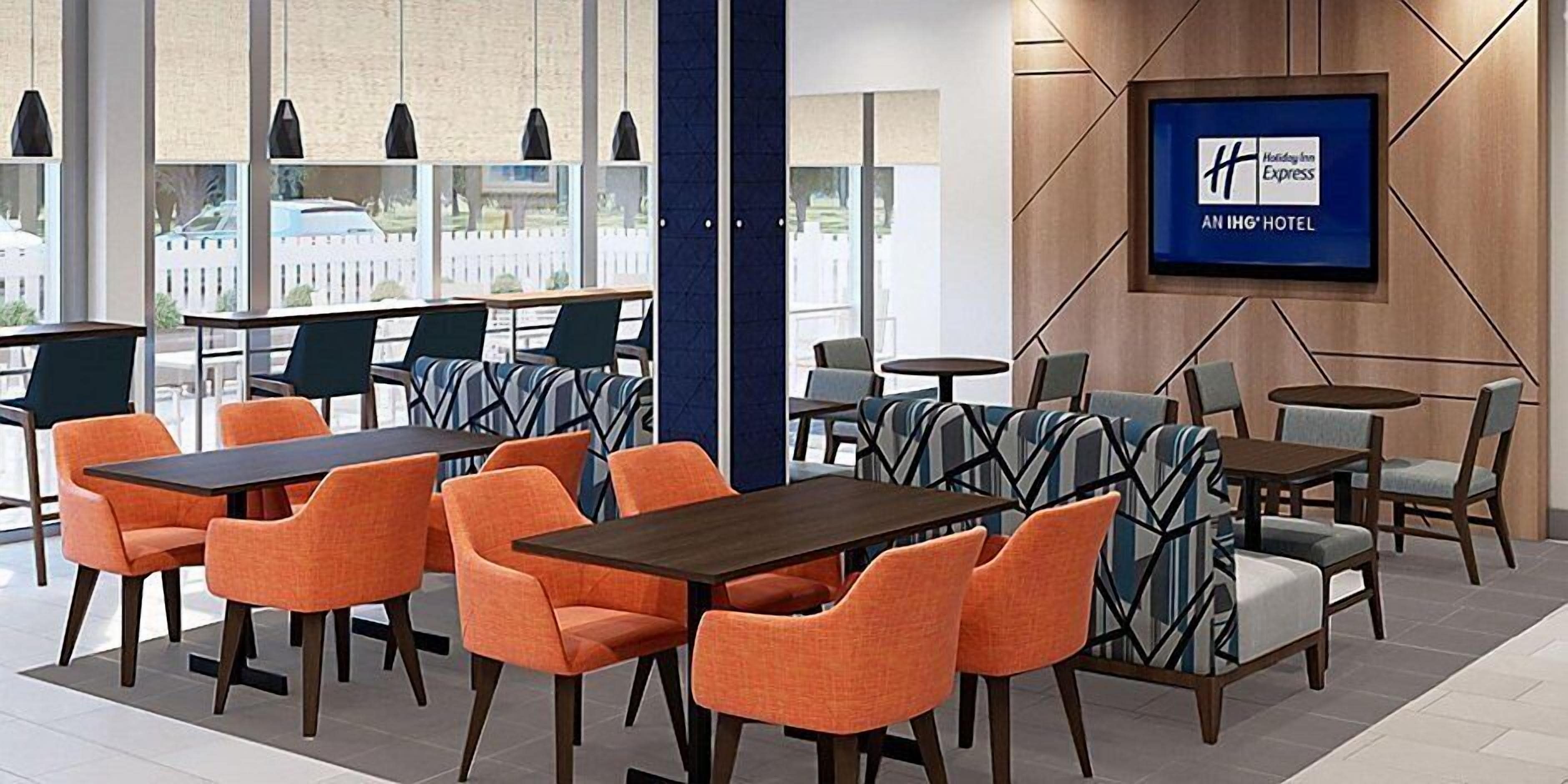 Un hall d’hôtel moderne avec des sièges orange et gris, un grand écran de télévision affichant le nom de l’hôtel et une vue sur l’extérieur grâce à de grandes fenêtres.