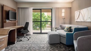 Habitación de hotel con escritorio, televisión, sofá, reposapiés y un ventanal con vistas a zonas verdes.