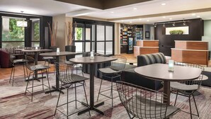 Un moderno vestíbulo de hotel con mesas redondas, taburetes de bar y una zona de estar con sofás. Amplias ventanas permiten la entrada de luz natural, un mostrador de recepción y un refrigerador con bebidas.