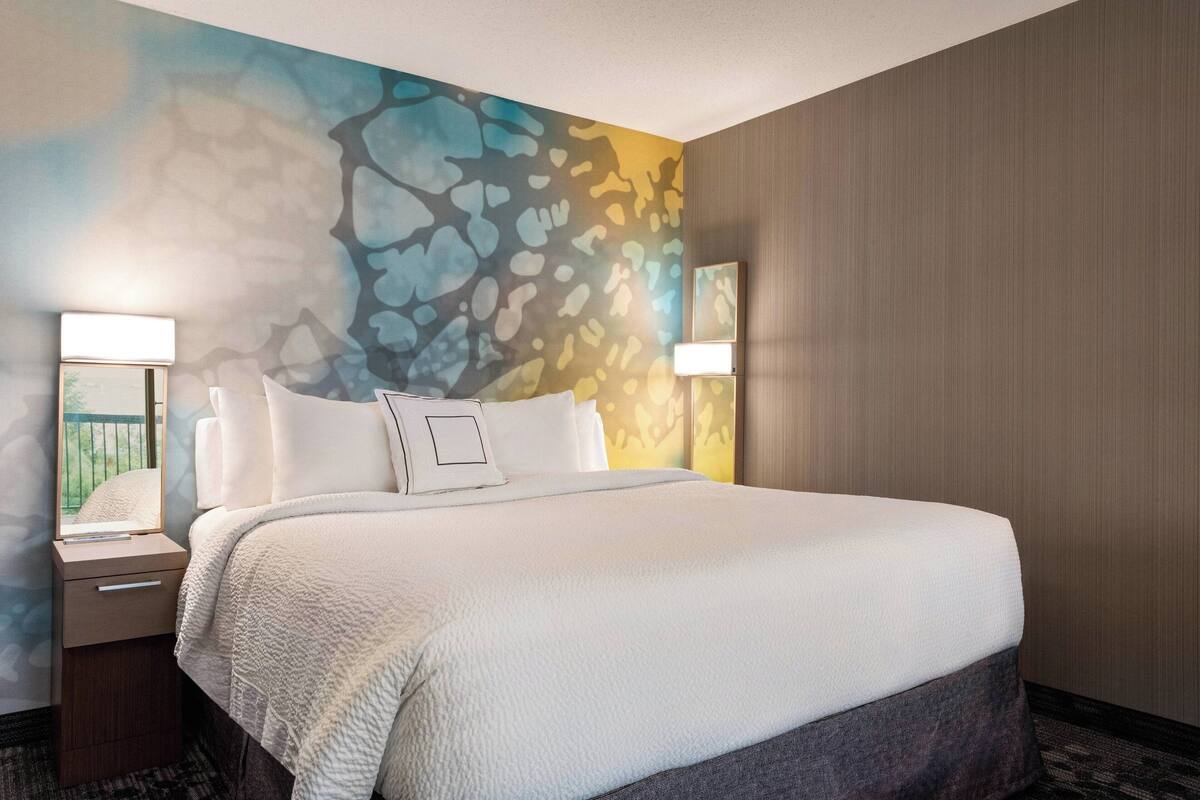 Una habitación de hotel con una cama grande, dos lámparas de noche, un mesita de noche y una pared con un mural abstracto colorido.