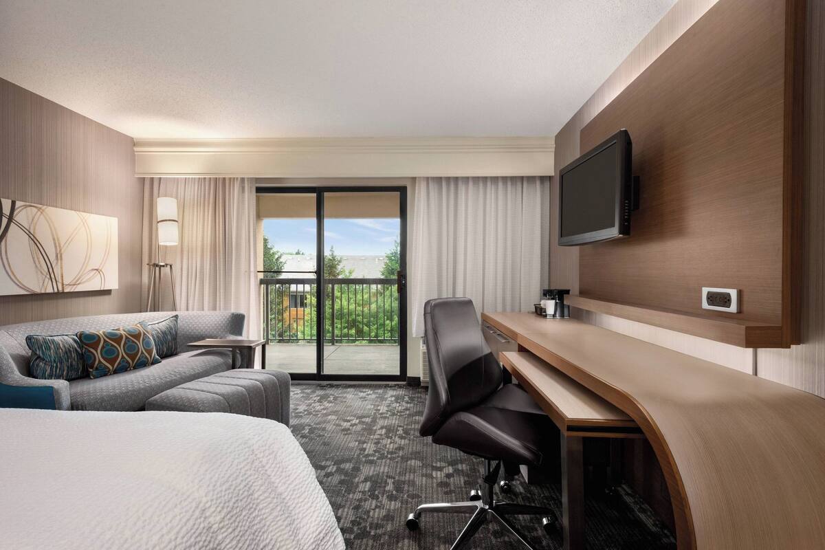Habitación de hotel con cama, escritorio, silla, televisor y balcón con vistas.