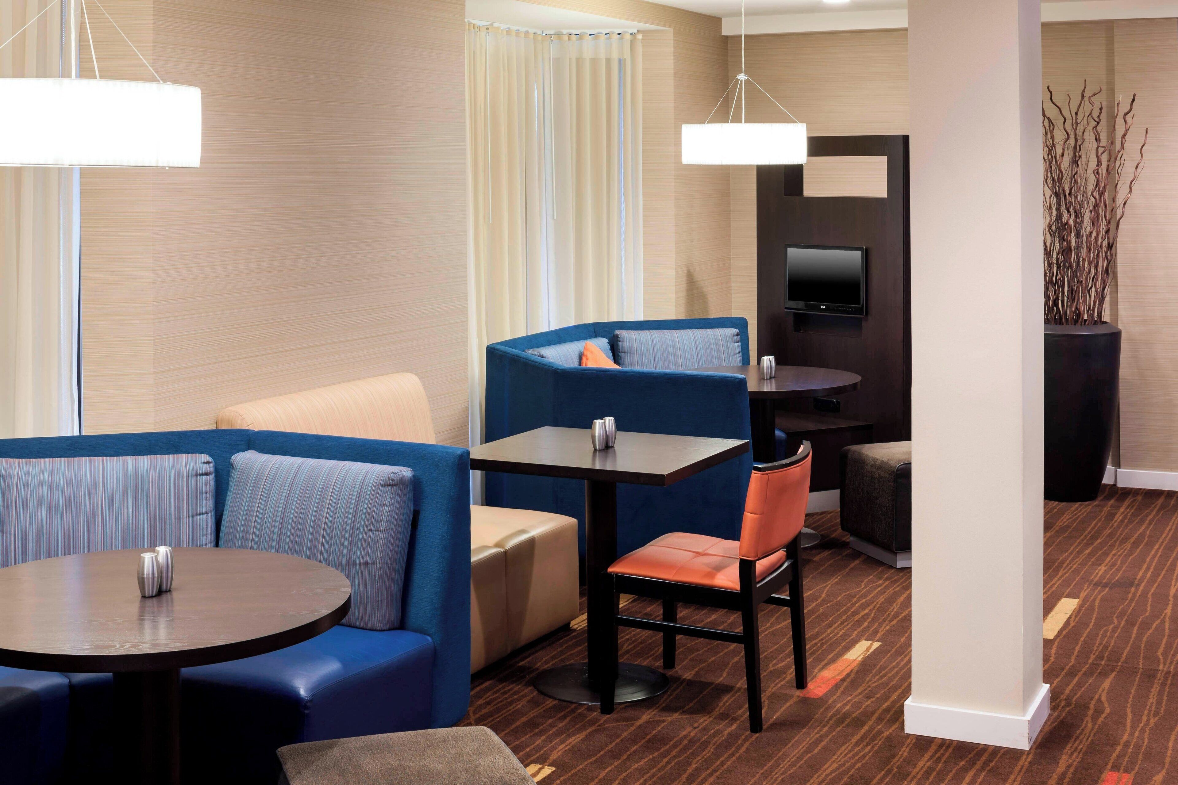 A hotel room with a sectional sofa, a round table, a small table, a television, and a decorative plant.