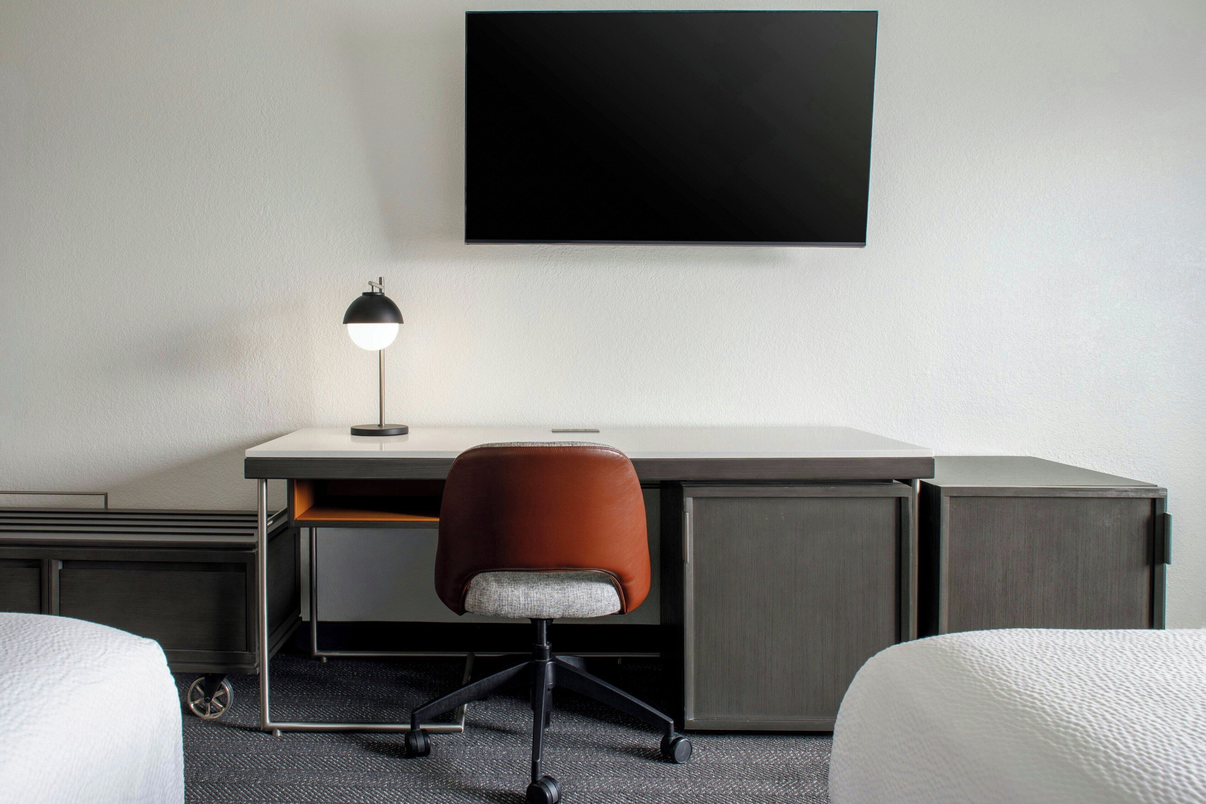 A hotel room with a desk, a chair, a TV mounted on the wall, and two beds.