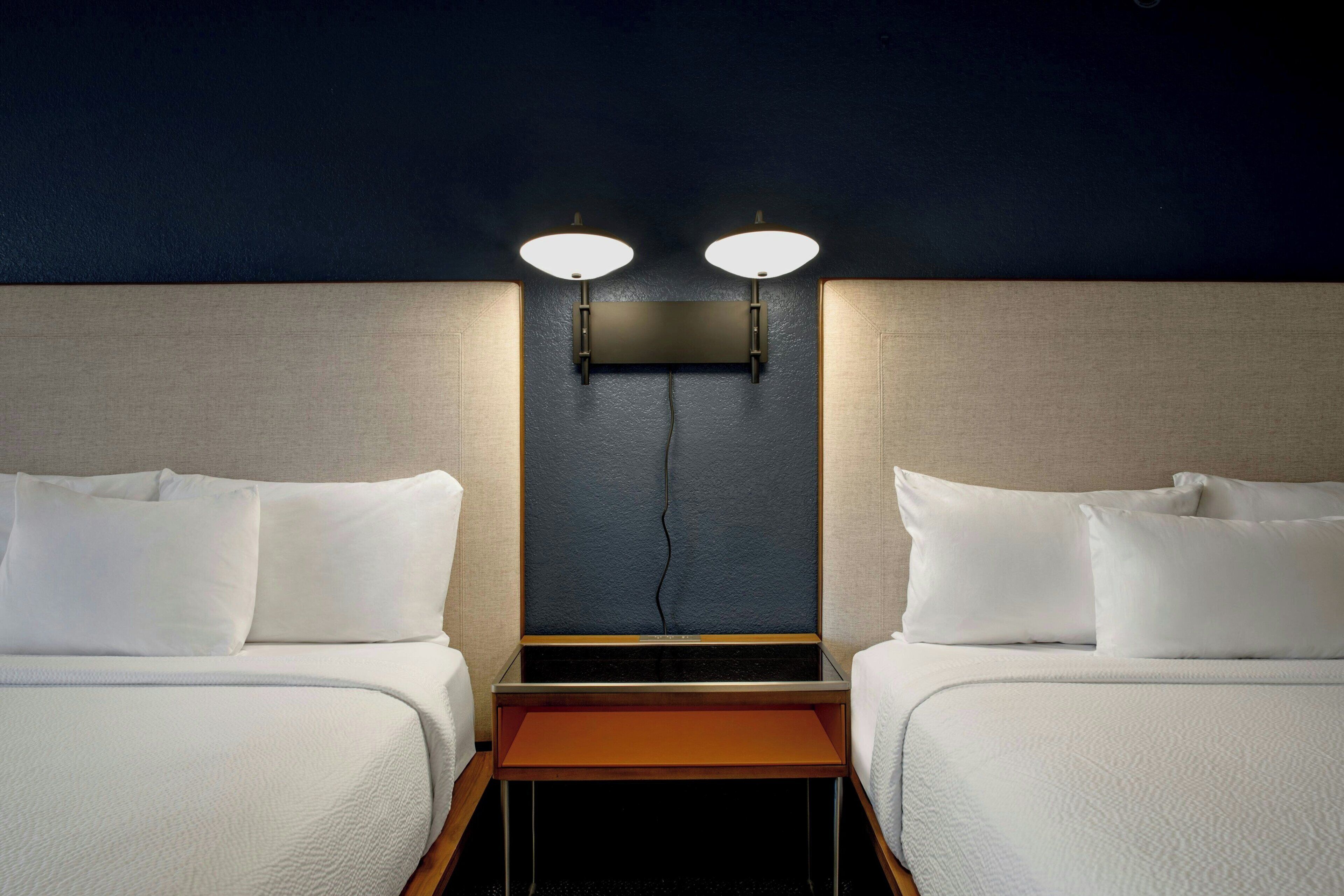 A hotel room with two beds, a nightstand, and a wall-mounted light fixture.