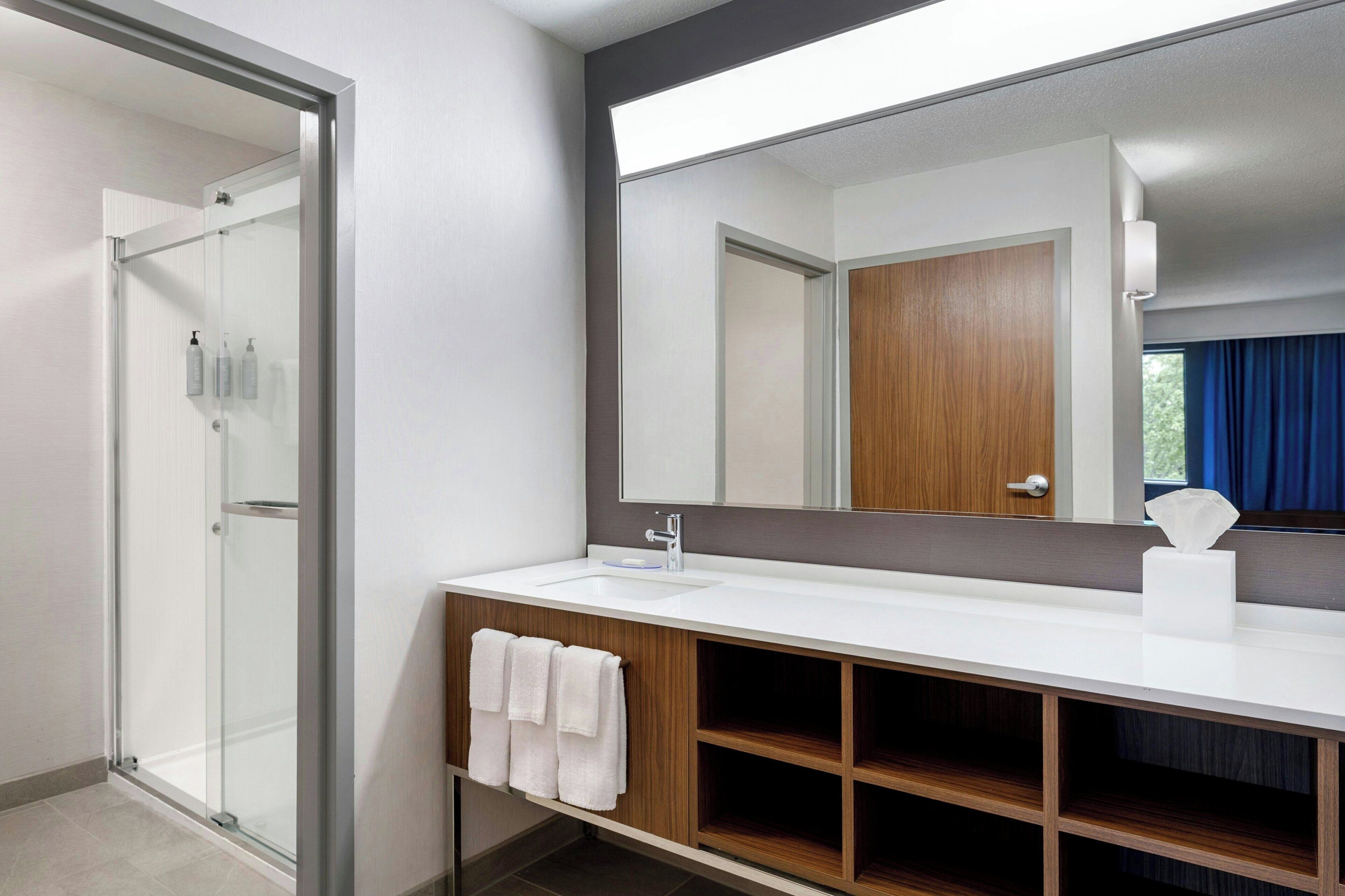 Een moderne badkamer met een douchecabine van glas, een witte wastafel met houten kastje, en een grote spiegel die een deur en een raam met blauwe gordijnen weerspiegelt.