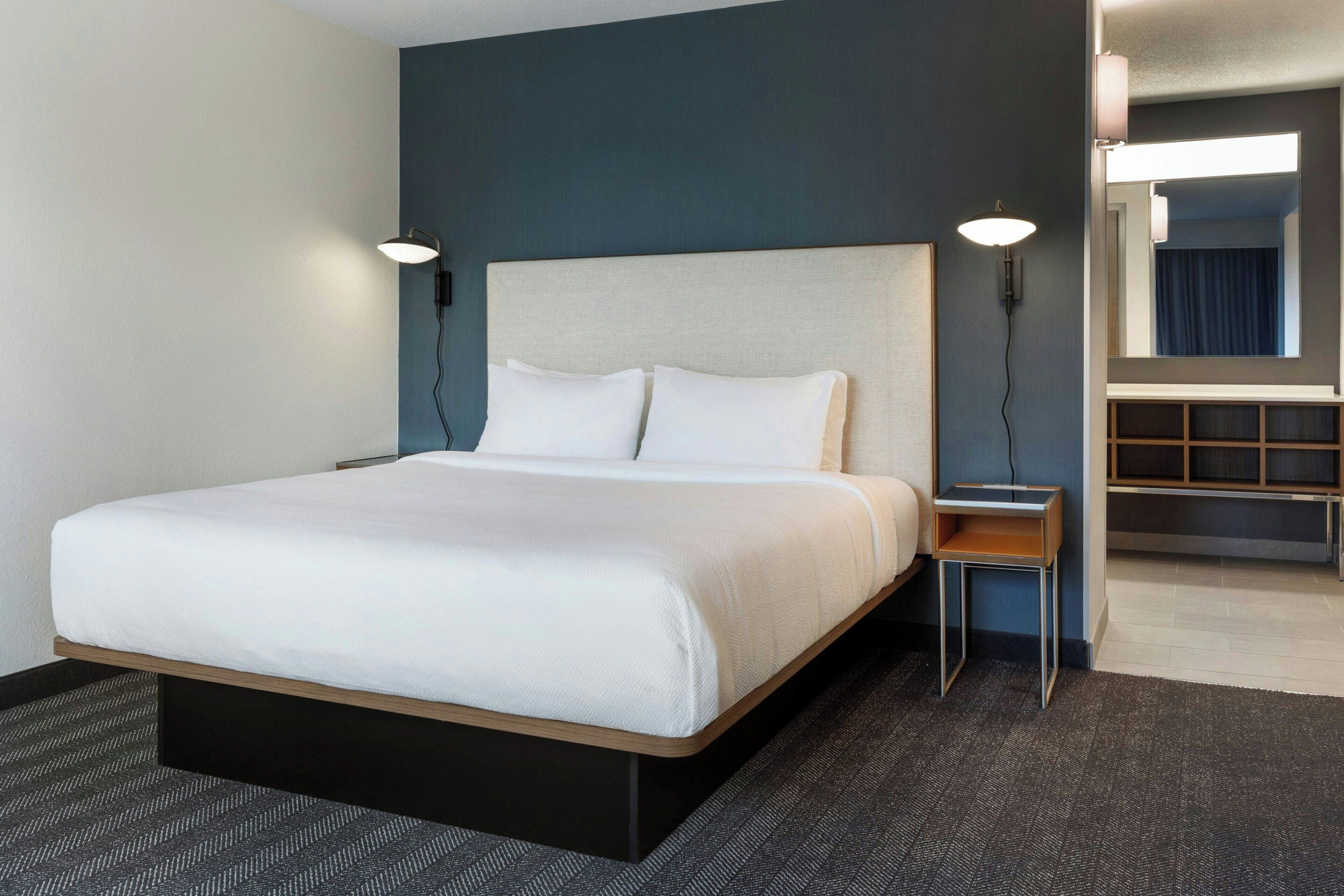A hotel room with a large bed, a nightstand, a wall-mounted lamp, and a mirror.