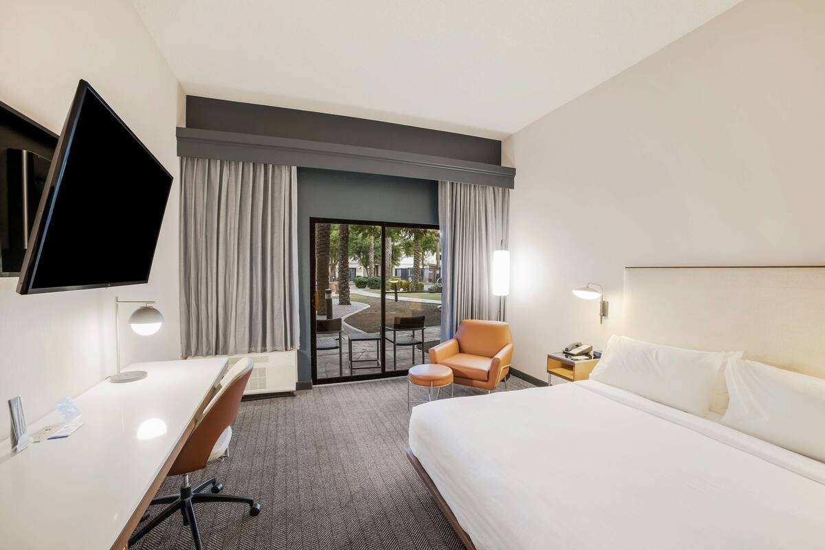 A hotel room with a bed, desk, chair, TV, and a view of the outdoors.