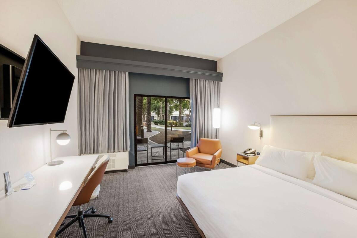 A hotel room with a bed, desk, chair, TV, and a view of the outdoor area.