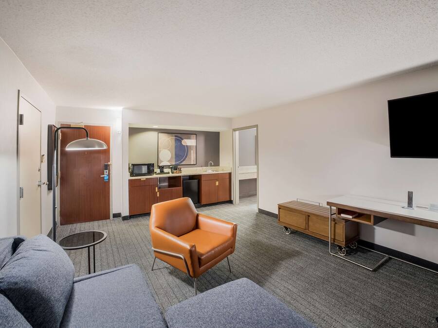 A hotel room with a sofa, a chair, a desk with a flat-screen TV, a kitchenette, and a door leading to another room.