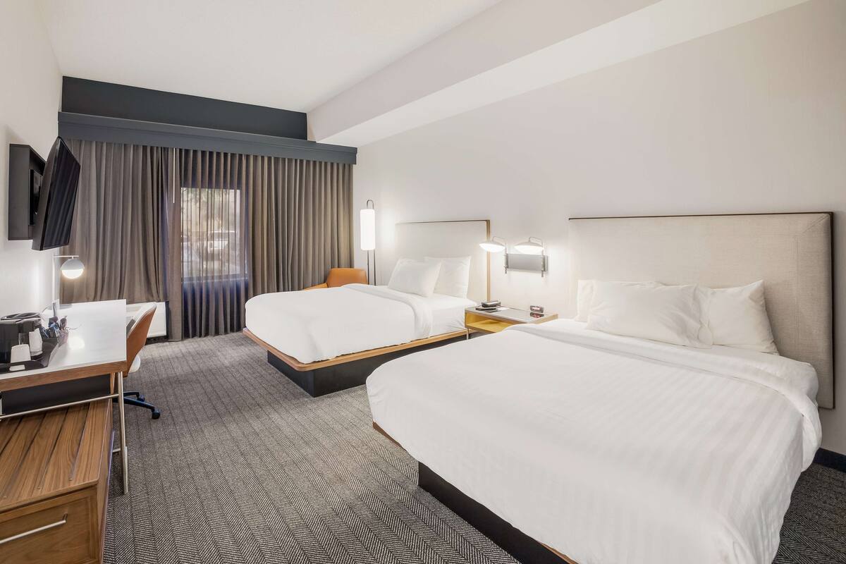 A hotel room with two beds, a desk, a chair, a TV, and a window with curtains.
