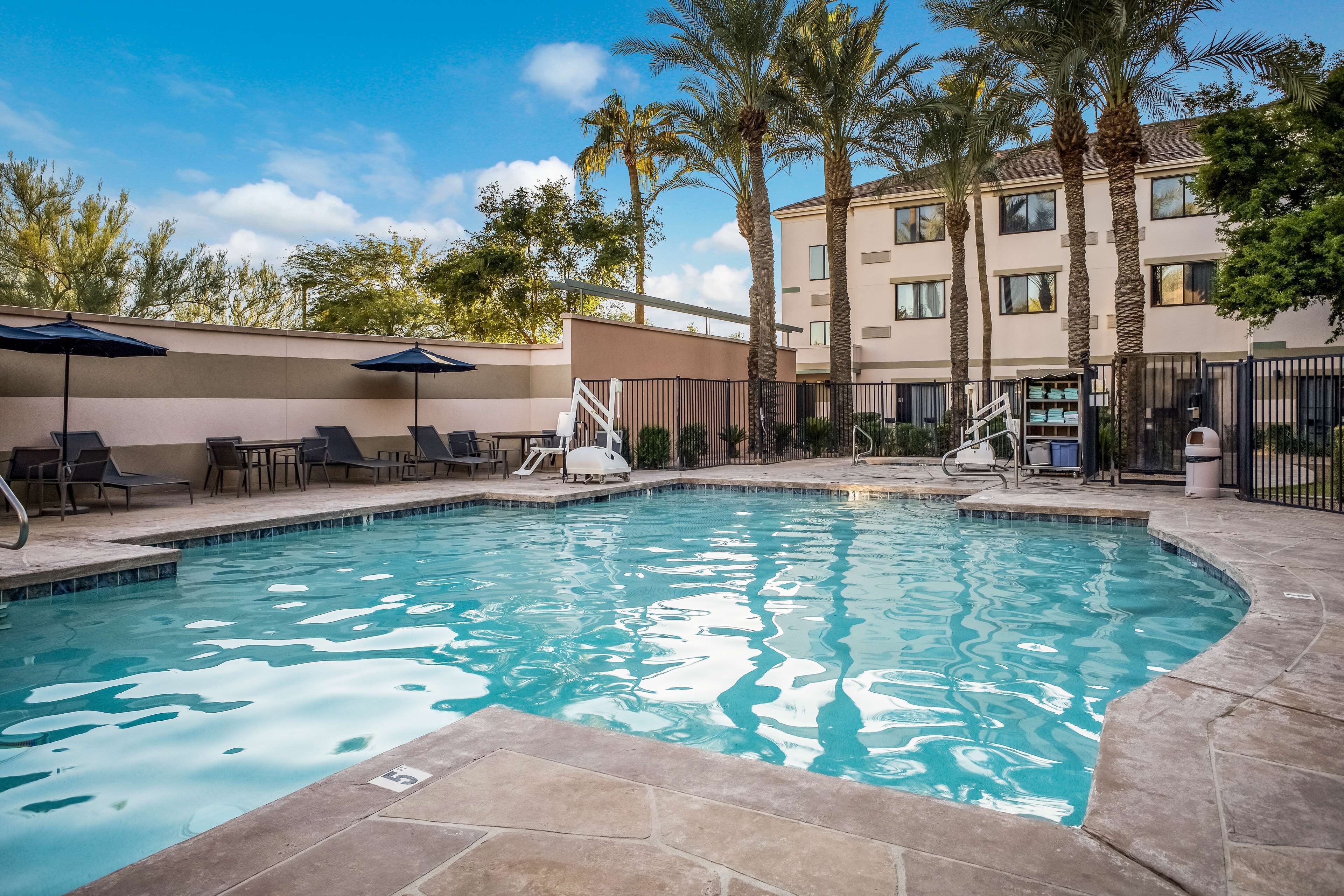A swimming pool with lounge chairs and umbrellas, surrounded by palm trees and a building with multiple windows.