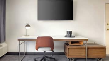 Habitación de hotel con televisor de pantalla plana montado en la pared, escritorio con silla, lámpara y un pequeño armario de madera con ruedas.