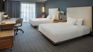 A hotel room with two beds, a desk, a chair, a TV, and a large window with curtains.