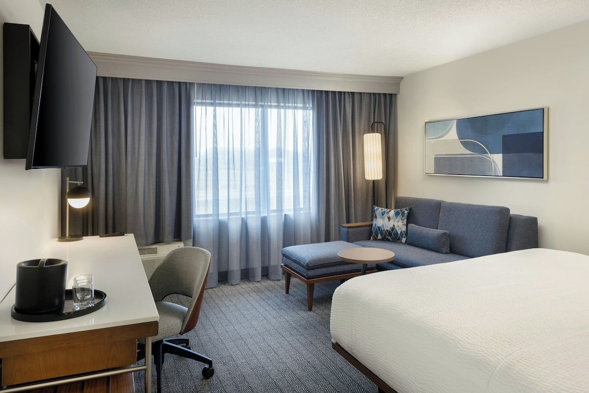 A hotel room with a bed, a desk with a chair, a sofa, a small table, a TV, and a window with curtains.