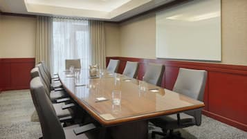 A conference room with a long table, multiple chairs, a projector screen, and a large window with curtains.