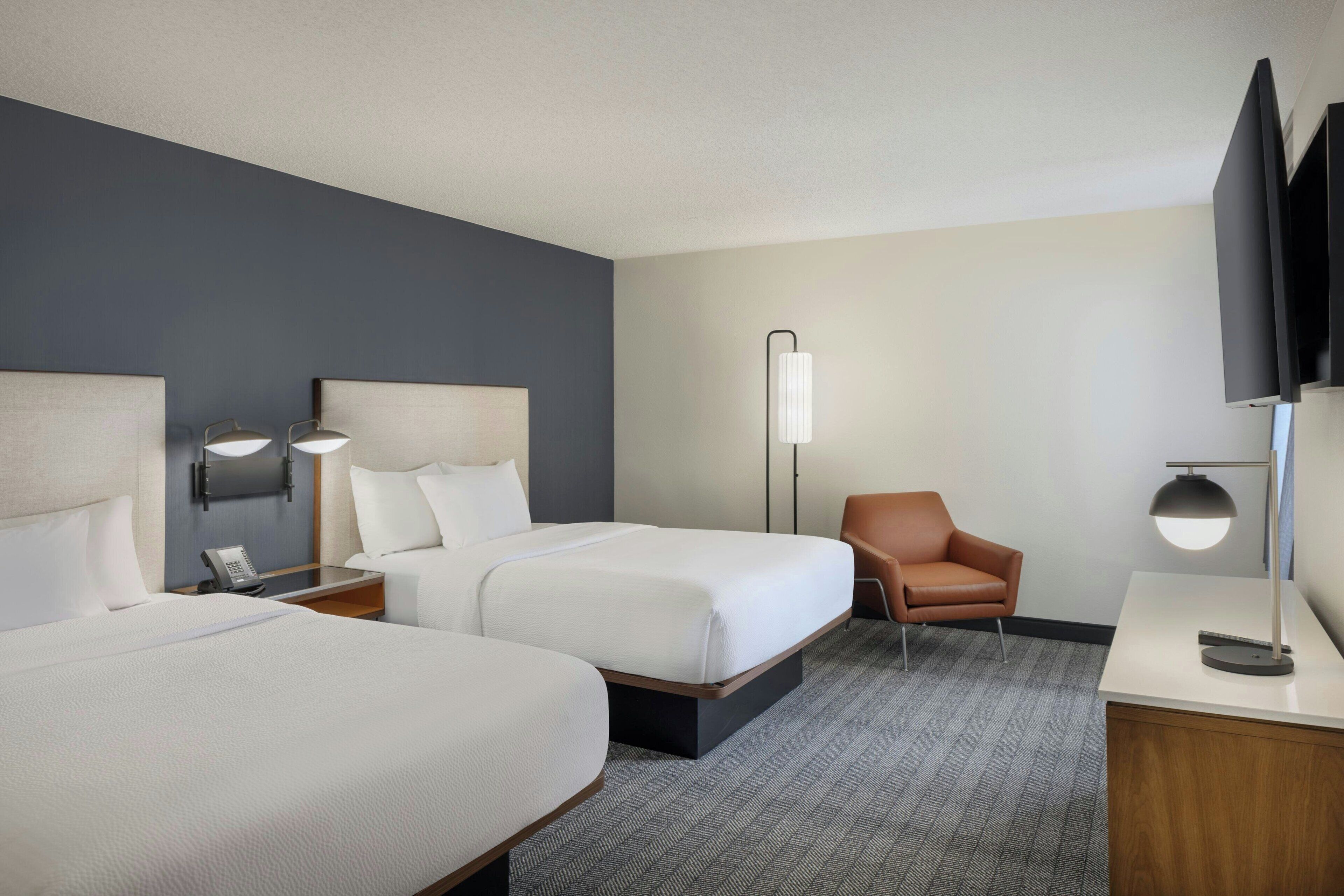 A hotel room with two beds, a desk, a chair, a TV, and a lamp.
