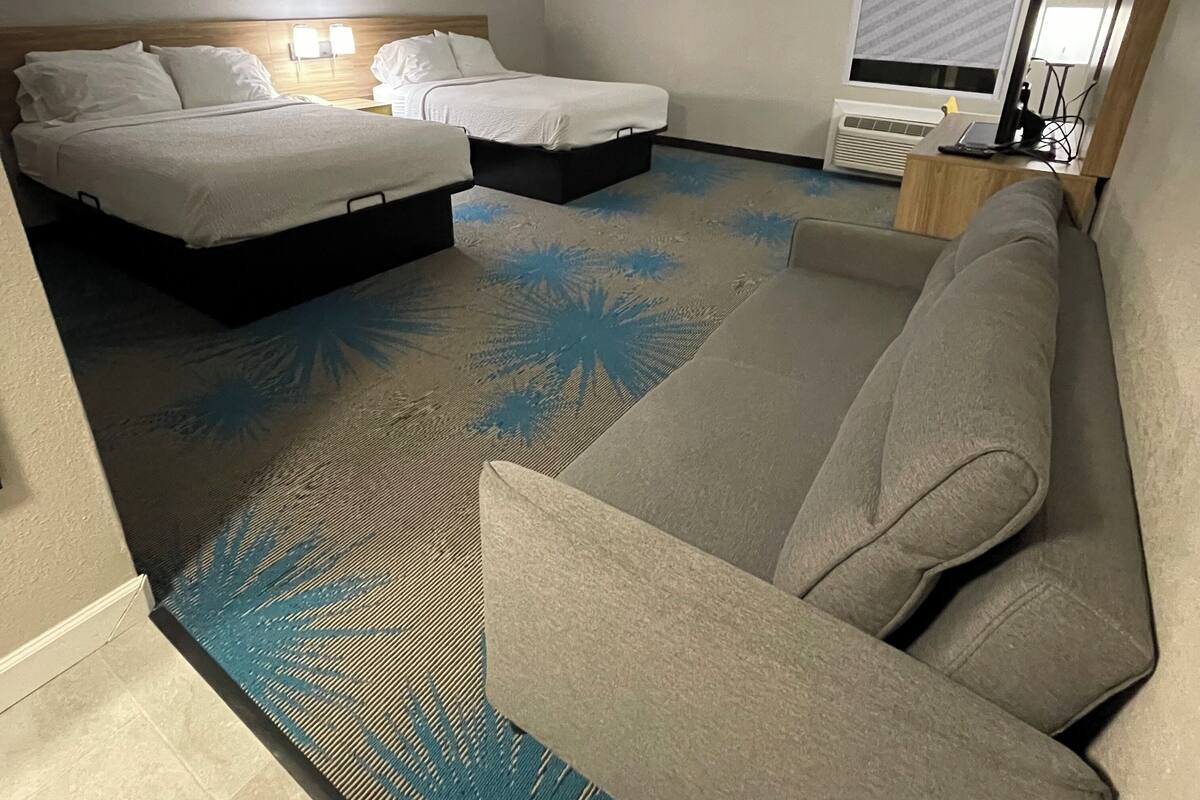 Ein Hotelzimmer mit zwei Betten, einem Sofa, einem gemusterten Teppich und einem Fenster mit Jalousien.